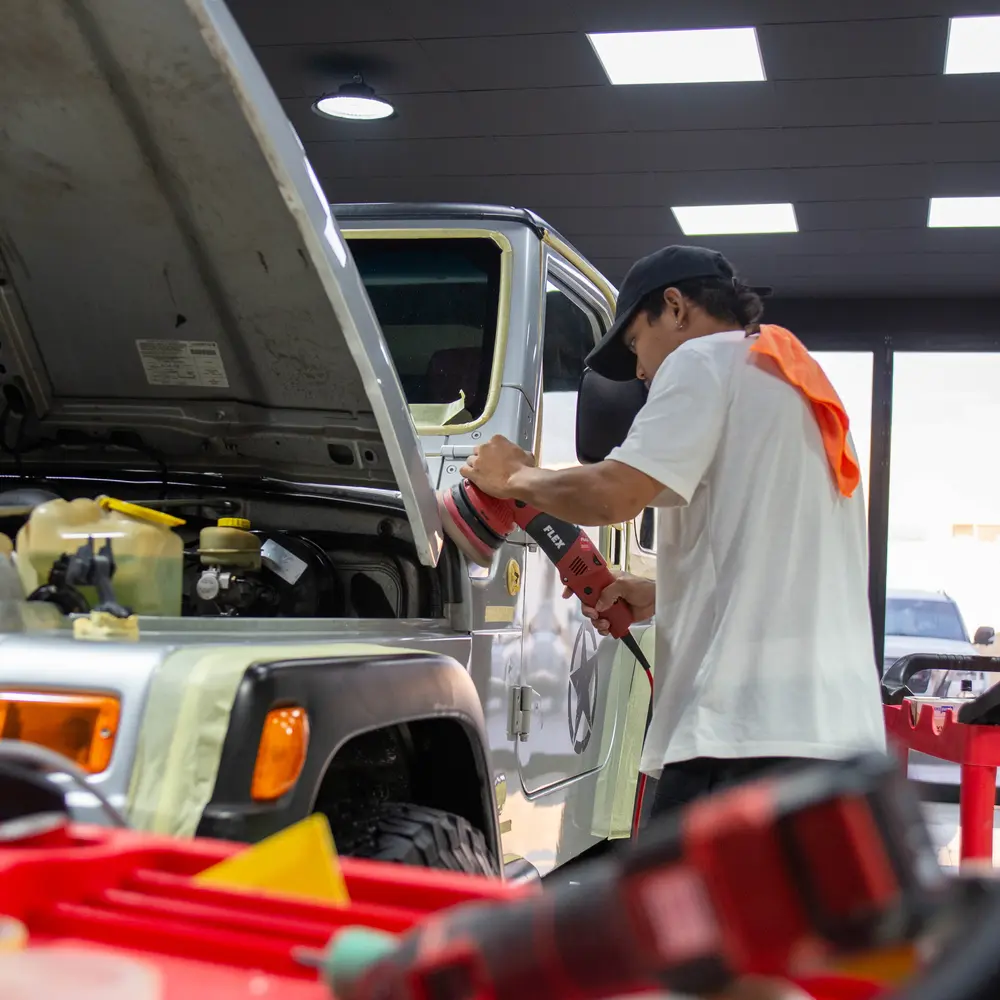 Detailer buffing a grayish-blue Jeep Wrangler with a buffing tool, with a tool cart in the foreground.